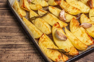 Baked pieces of spiced potato in a baking pan. Fragrant rosemary, garlic, sea salt, oil. Vegan healthy food concept. Vintage wooden boards background, close up