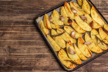 Baked pieces of spiced potato in a baking pan. Fragrant rosemary, garlic, sea salt, oil. Vegan healthy food concept. Vintage wooden boards background, top view