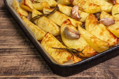 Baked pieces of spiced potato in a baking pan. Fragrant rosemary, garlic, sea salt, oil. Vegan healthy food concept. Vintage wooden boards background, close up