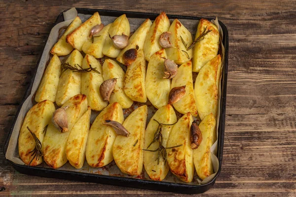 Baked pieces of spiced potato in a baking pan. Fragrant rosemary, garlic, sea salt, oil. Vegan healthy food concept. Vintage wooden boards background, copy space