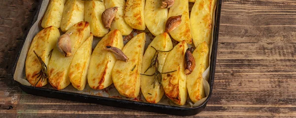 Baked pieces of spiced potato in a baking pan. Fragrant rosemary, garlic, sea salt, oil. Vegan healthy food concept. Vintage wooden boards background, banner format