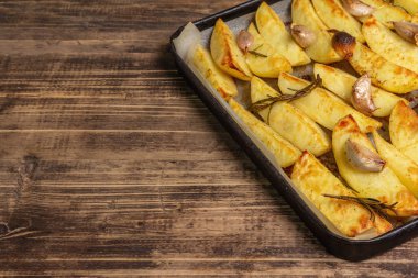 Baked pieces of spiced potato in a baking pan. Fragrant rosemary, garlic, sea salt, oil. Vegan healthy food concept. Vintage wooden boards background, copy space
