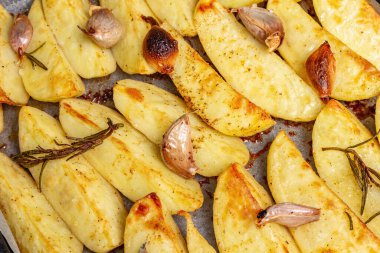 Baked pieces of spiced potato in a baking pan. Fragrant rosemary, garlic, sea salt, oil. Vegan healthy food concept. Vintage wooden boards background, close up