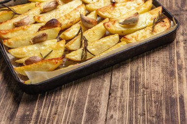 Baked pieces of spiced potato in a baking pan. Fragrant rosemary, garlic, sea salt, oil. Vegan healthy food concept. Vintage wooden boards background, close up