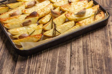 Baked pieces of spiced potato in a baking pan. Fragrant rosemary, garlic, sea salt, oil. Vegan healthy food concept. Vintage wooden boards background, close up