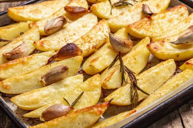 Baked pieces of spiced potato in a baking pan. Fragrant rosemary, garlic, sea salt, oil. Vegan healthy food concept. Vintage wooden boards background, close up