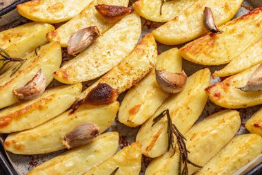 Baked pieces of spiced potato in a baking pan. Fragrant rosemary, garlic, sea salt, oil. Vegan healthy food concept. Vintage wooden boards background, close up