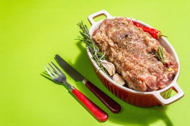 Sarımsaklı çiğ domuz rulosu ve seramik kızartma kabında havuç. Taze et, çatal bıçak, baharat, biberiye, sarımsak. Açık yeşil arkaplan, modern sert ışık, koyu gölge, kopyalama alanı