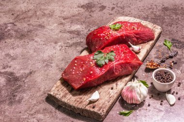 Tahta kesme tahtasında çiğ biftek. Modaya uygun sert ışık, karanlık gölge. Baharatlar, taze sarımsak ve maydanoz. Taş beton arkaplan, kopyalama alanı