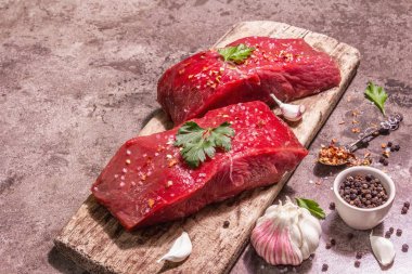 Tahta kesme tahtasında çiğ biftek. Modaya uygun sert ışık, karanlık gölge. Baharatlar, taze sarımsak ve maydanoz. Taş beton arkaplan, kopyalama alanı