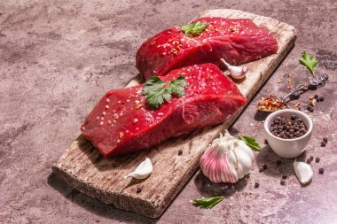 Tahta kesme tahtasında çiğ biftek. Modaya uygun sert ışık, karanlık gölge. Baharatlar, taze sarımsak ve maydanoz. Taş beton arkaplan, kopyalama alanı