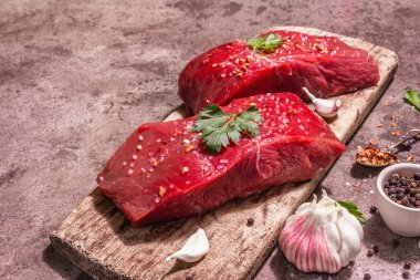 Tahta kesme tahtasında çiğ biftek. Modaya uygun sert ışık, karanlık gölge. Baharatlar, taze sarımsak ve maydanoz. Taş beton arka plan, kapat