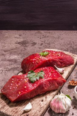 Tahta kesme tahtasında çiğ biftek. Modaya uygun sert ışık, karanlık gölge. Baharatlar, taze sarımsak ve maydanoz. Taş beton arkaplan, kopyalama alanı