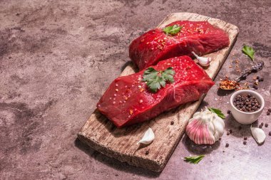 Tahta kesme tahtasında çiğ biftek. Modaya uygun sert ışık, karanlık gölge. Baharatlar, taze sarımsak ve maydanoz. Taş beton arkaplan, kopyalama alanı