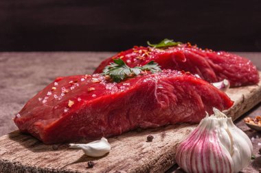 Tahta kesme tahtasında çiğ biftek. Modaya uygun sert ışık, karanlık gölge. Baharatlar, taze sarımsak ve maydanoz. Taş beton arka plan, kapat