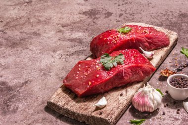 Tahta kesme tahtasında çiğ biftek. Modaya uygun sert ışık, karanlık gölge. Baharatlar, taze sarımsak ve maydanoz. Taş beton arkaplan, kopyalama alanı