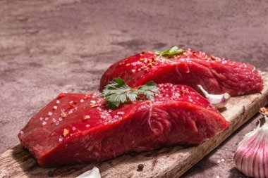 Tahta kesme tahtasında çiğ biftek. Modaya uygun sert ışık, karanlık gölge. Baharatlar, taze sarımsak ve maydanoz. Taş beton arka plan, kapat