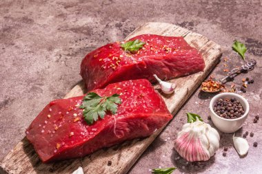 Tahta kesme tahtasında çiğ biftek. Modaya uygun sert ışık, karanlık gölge. Baharatlar, taze sarımsak ve maydanoz. Taş beton arkaplan, kopyalama alanı