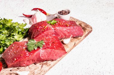 Tahta kesme tahtasında çiğ biftek. Modaya uygun sert ışık, karanlık gölge. Baharatlar, acı biber, taze sarımsak ve maydanoz. Hafif alçı arkaplan, kopyalama alanı