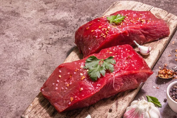 Tahta kesme tahtasında çiğ biftek. Modaya uygun sert ışık, karanlık gölge. Baharatlar, taze sarımsak ve maydanoz. Taş beton arkaplan, kopyalama alanı