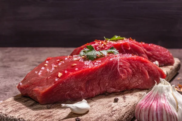 Tahta kesme tahtasında çiğ biftek. Modaya uygun sert ışık, karanlık gölge. Baharatlar, taze sarımsak ve maydanoz. Taş beton arka plan, kapat