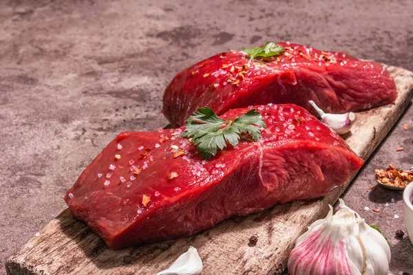 Tahta kesme tahtasında çiğ biftek. Modaya uygun sert ışık, karanlık gölge. Baharatlar, taze sarımsak ve maydanoz. Taş beton arka plan, kapat