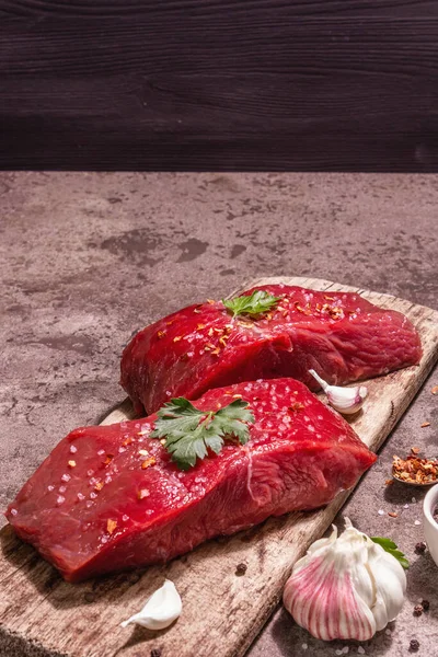 Tahta kesme tahtasında çiğ biftek. Modaya uygun sert ışık, karanlık gölge. Baharatlar, taze sarımsak ve maydanoz. Taş beton arkaplan, kopyalama alanı