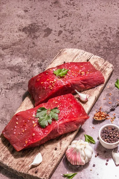 Tahta kesme tahtasında çiğ biftek. Modaya uygun sert ışık, karanlık gölge. Baharatlar, taze sarımsak ve maydanoz. Taş beton zemin, üst görünüm