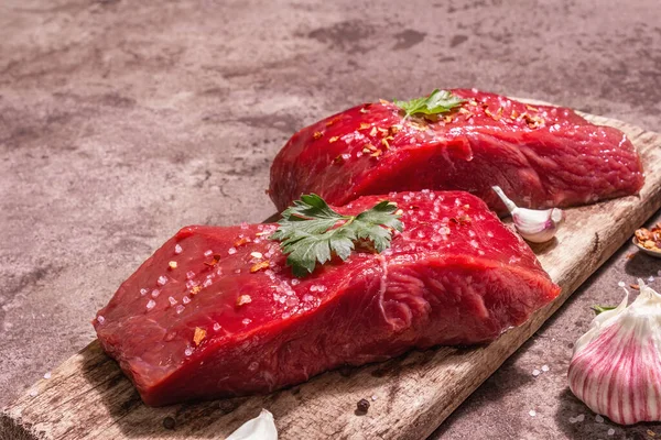 Tahta kesme tahtasında çiğ biftek. Modaya uygun sert ışık, karanlık gölge. Baharatlar, taze sarımsak ve maydanoz. Taş beton arkaplan, kopyalama alanı