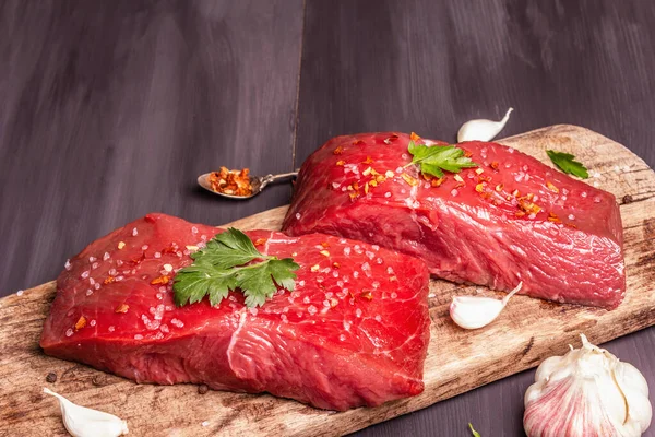 Tahta kesme tahtasında çiğ biftek. Baharatlar, taze sarımsak ve maydanoz. Siyah tahta kaplama arka plan, kapat