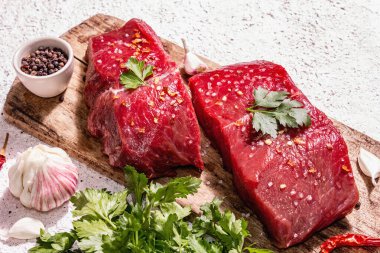 Tahta kesme tahtasında çiğ biftek. Modaya uygun sert ışık, karanlık gölge. Baharatlar, acı biber, taze sarımsak ve maydanoz. Hafif alçı arkaplan, kapat