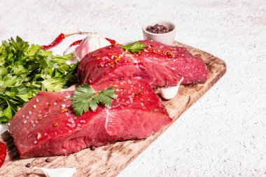 Tahta kesme tahtasında çiğ biftek. Modaya uygun sert ışık, karanlık gölge. Baharatlar, acı biber, taze sarımsak ve maydanoz. Hafif alçı arkaplan, kopyalama alanı