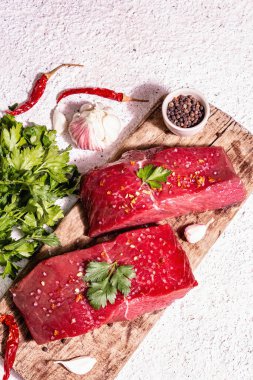 Tahta kesme tahtasında çiğ biftek. Modaya uygun sert ışık, karanlık gölge. Baharatlar, acı biber, taze sarımsak ve maydanoz. Hafif alçı arkaplan, üst görünüm