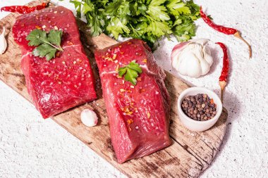 Tahta kesme tahtasında çiğ biftek. Modaya uygun sert ışık, karanlık gölge. Baharatlar, acı biber, taze sarımsak ve maydanoz. Hafif alçı arkaplan, kapat