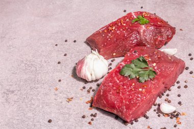 Gri taşlı arka planda çiğ biftek. Modaya uygun sert ışık, karanlık gölge. Baharatlar, acı biber, dağınık siyah biber, taze sarımsak ve maydanoz, kopyalama alanı.