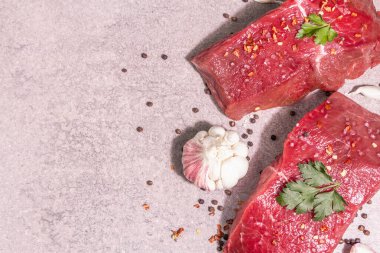 Gri taşlı arka planda çiğ biftek. Modaya uygun sert ışık, karanlık gölge. Baharatlar, acı biber, dağınık siyah biber, taze sarımsak ve maydanoz.