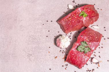 Gri taşlı arka planda çiğ biftek. Modaya uygun sert ışık, karanlık gölge. Baharatlar, acı biber, dağınık siyah biber, taze sarımsak ve maydanoz.