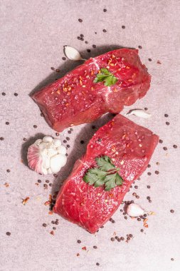 Gri taşlı arka planda çiğ biftek. Modaya uygun sert ışık, karanlık gölge. Baharatlar, acı biber, dağınık siyah biber, taze sarımsak ve maydanoz.