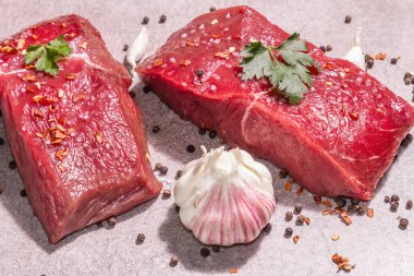 Gri taşlı arka planda çiğ biftek. Modaya uygun sert ışık, karanlık gölge. Baharatlar, acı biber, dağınık karabiber, taze sarımsak ve maydanoz.