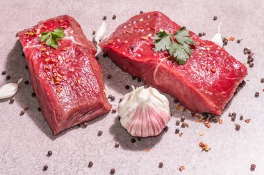 Gri taşlı arka planda çiğ biftek. Modaya uygun sert ışık, karanlık gölge. Baharatlar, acı biber, dağınık karabiber, taze sarımsak ve maydanoz.