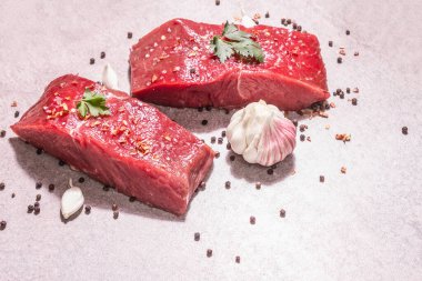 Gri taşlı arka planda çiğ biftek. Modaya uygun sert ışık, karanlık gölge. Baharatlar, acı biber, dağınık siyah biber, taze sarımsak ve maydanoz, kopyalama alanı.