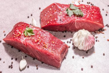 Gri taşlı arka planda çiğ biftek. Modaya uygun sert ışık, karanlık gölge. Baharatlar, acı biber, dağınık karabiber, taze sarımsak ve maydanoz.