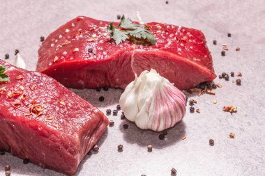 Gri taşlı arka planda çiğ biftek. Modaya uygun sert ışık, karanlık gölge. Baharatlar, acı biber, dağınık karabiber, taze sarımsak ve maydanoz.