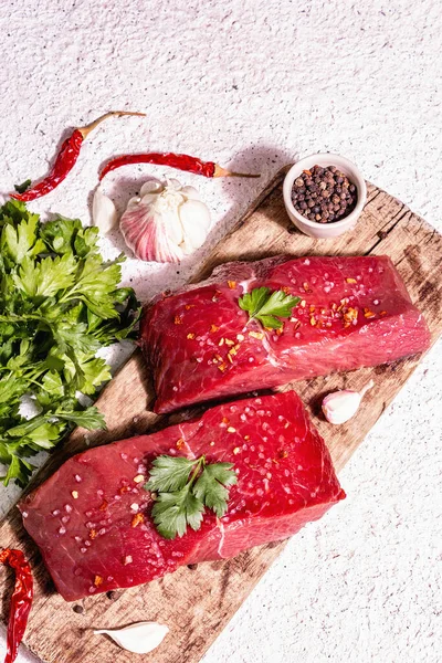 Tahta kesme tahtasında çiğ biftek. Modaya uygun sert ışık, karanlık gölge. Baharatlar, acı biber, taze sarımsak ve maydanoz. Hafif alçı arkaplan, üst görünüm