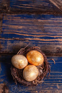 Hasır bir yuvadaki altın Paskalya yumurtaları. Geleneksel Paskalya sembolü, şenlikli dekor. Mavi renkli ahşap tahta arka plan, üst görünüm