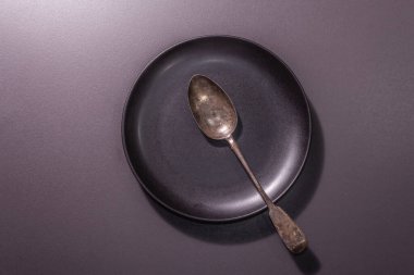Masa düzeneği olarak koyu seramik tabak ve klasik kaşık. Minimalist kavram, siyah taş beton zemin, sert ışık, koyu gölge, üst görüş
