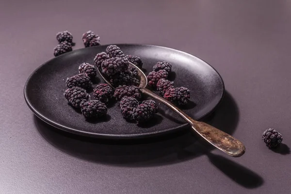 Seramik tabakta böğürtlen. Sağlıklı beslenme için dondurulmuş meyve. Minimalist konsept, siyah taş beton zemin, sert ışık, koyu gölge, düz uzanma