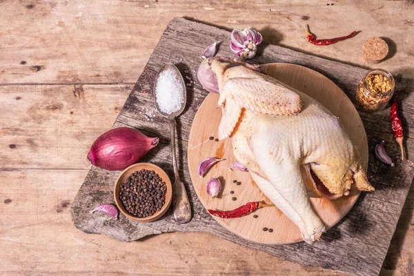 Raw whole chicken carcass for healthy food lifestyle. Free-range farm bird with red onion, garlic cloves, spices, and sea salt. A trendy hard light, dark shadow, vintage wooden table, top view