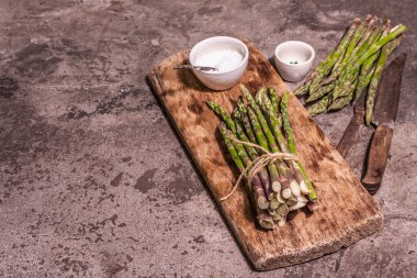 Bunch of fresh green asparagus on vintage wooden chopping board. Delicious ingredients ready for cooking healthy food. Modern hard light, dark shadow. Stone concrete background, copy space