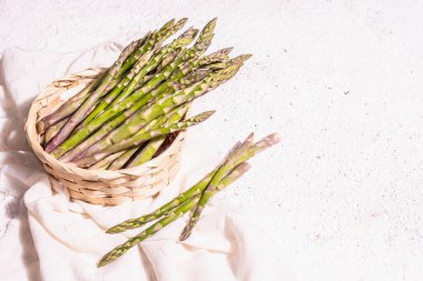 Ripe asparagus in a wicker basket. Fresh green ingredients ready for cooking healthy food. Modern hard light, dark shadow. White plaster background, copy space
